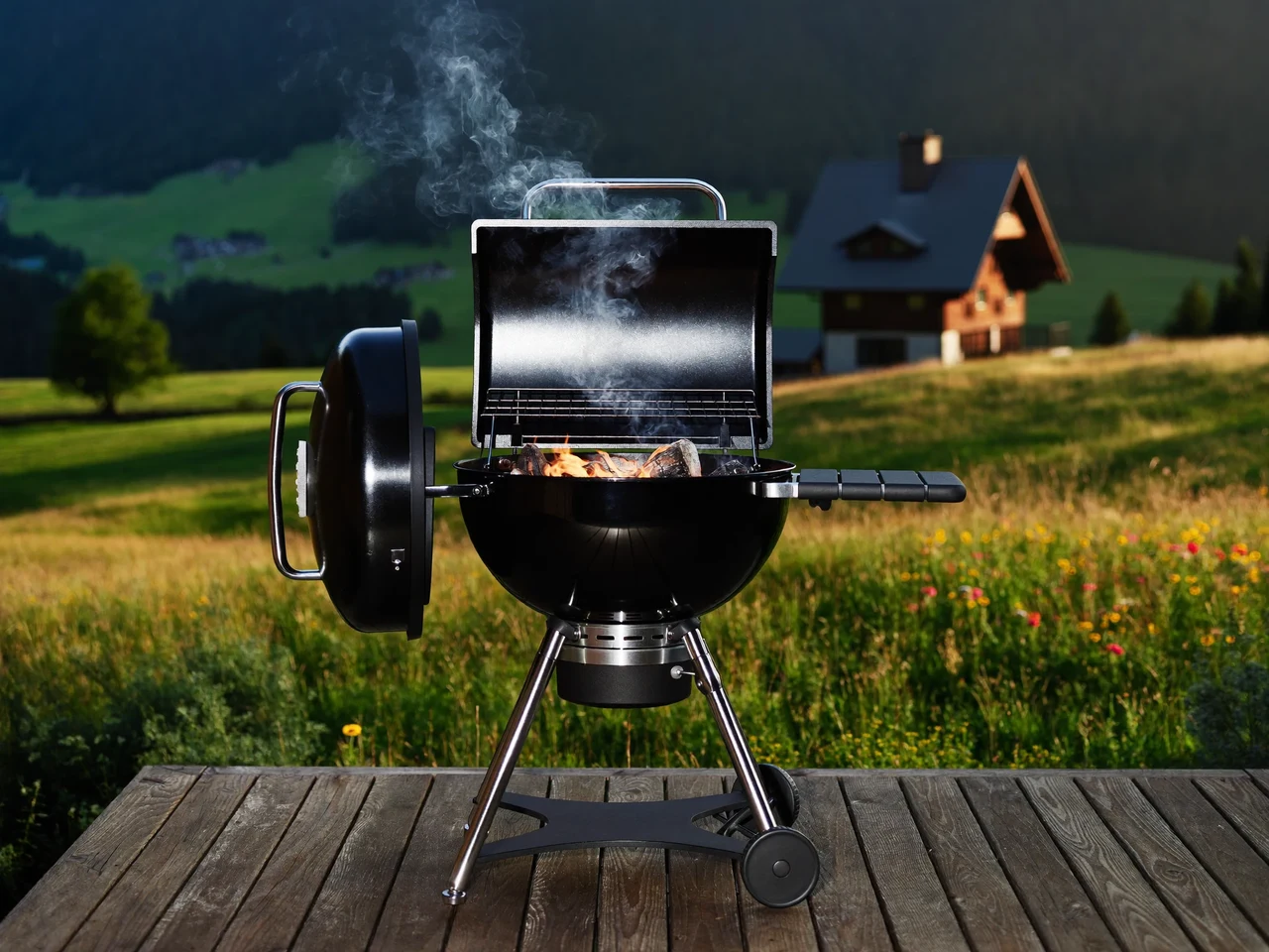 Brennender Holzkohlegrill auf einer Holzterrasse im goldenen Abendlicht vor Alpenwiese und Chalet